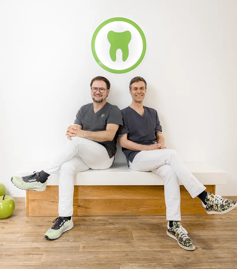 Roland und Stefan sitzen auf einer Bank, über ihnen ein grüner Zahn als Teil des Logos